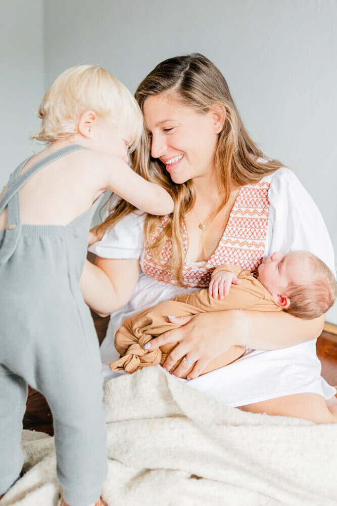 Mother smiles at her toddler while she holds a newborn at a session with a Boston Newborn Photographer