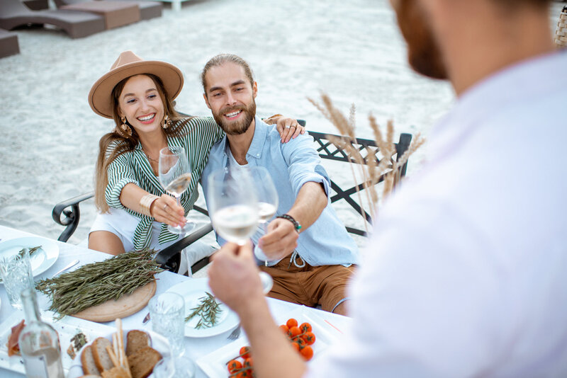 A smiling couple is sitting at an outdoor table on the beach, holding wine glasses for a toast. The table is set with food, herbs, and decor. Another person in the foreground is also raising a glass.