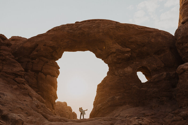 Couple stands under Turret Arch at sunrise in Arches National Park near Moab Utah
