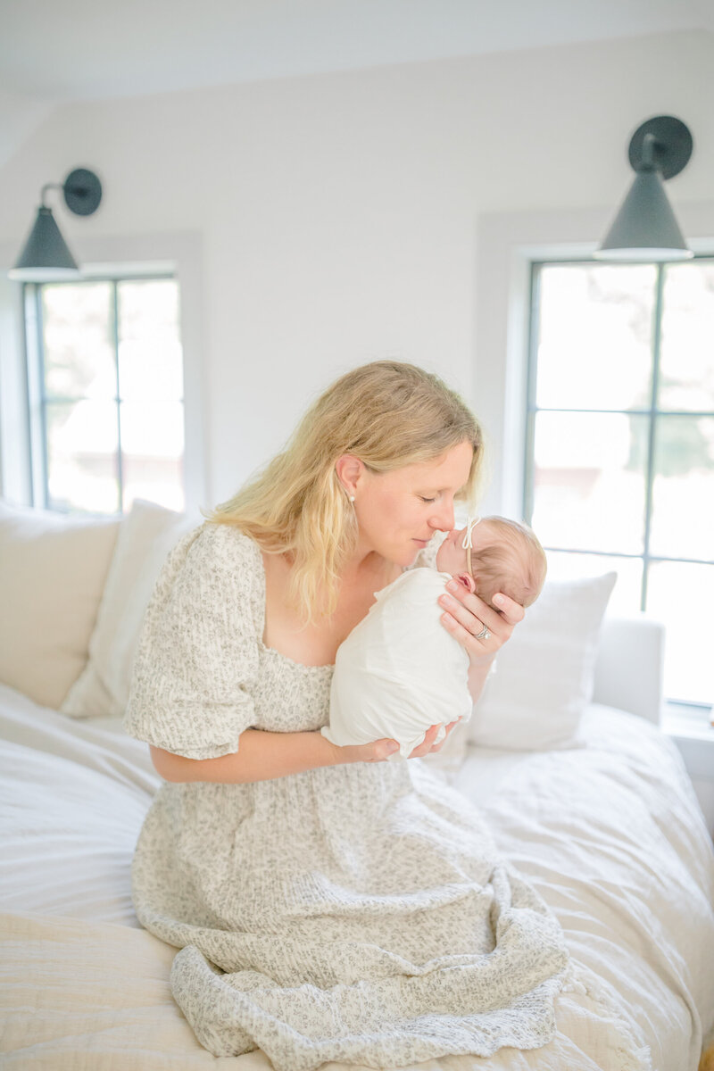 A new mother sits on her bed, cradling her swaddled newborn in a cozy in-home lifestyle session. She gently touches noses with her sleeping baby, sharing a tender moment filled with love and warmth