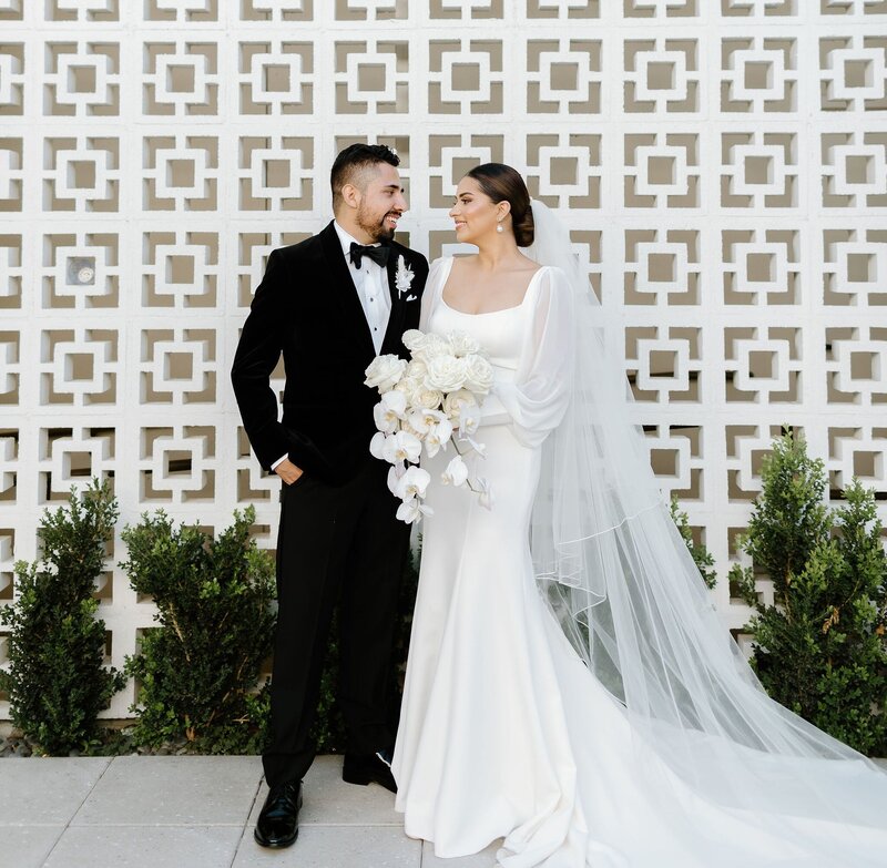 A bride and groom look lovingly at 
each other  in front of Hart and Main's breezeblock wall