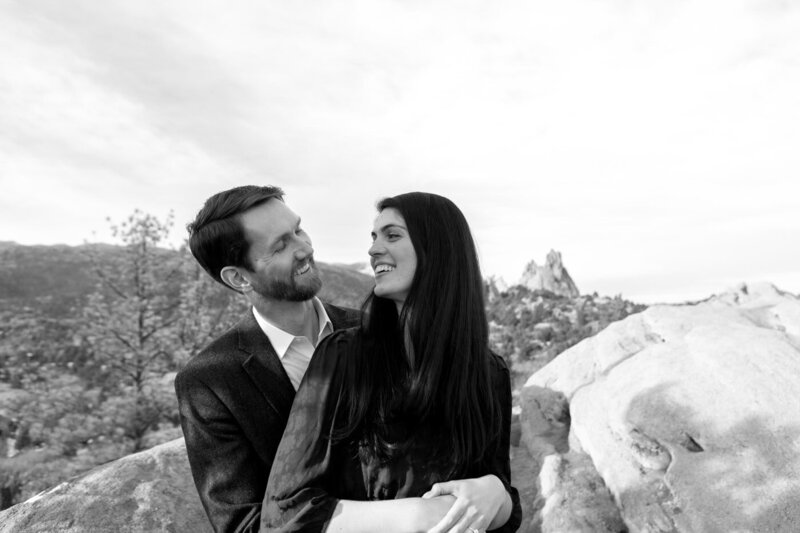 A couple with the man's arms wrapped around her waist turns to face each other by rocky outcroppings.
