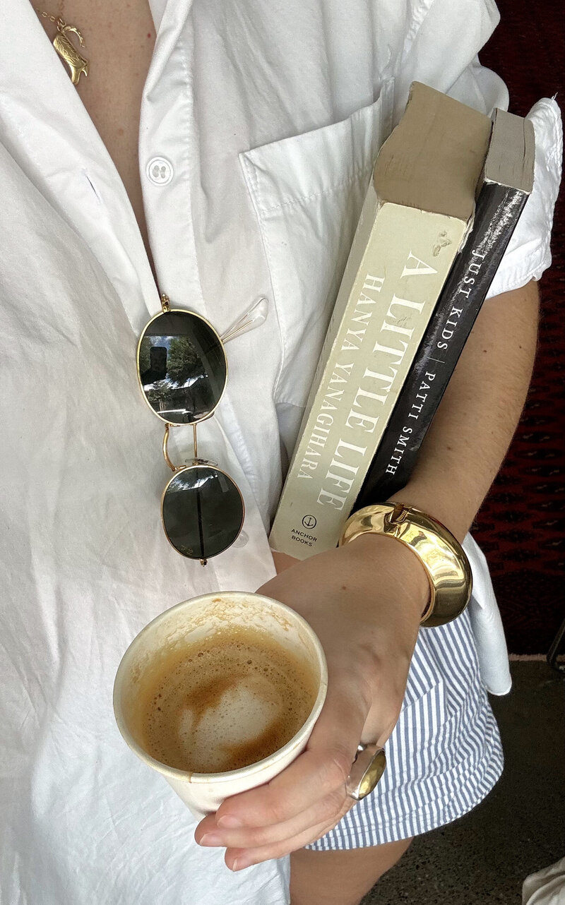 A person in a white shirt holds a coffee cup and two books, "A Little Life" and "Just Kids." Sunglasses hang from the pocket; gold bracelets are visible.