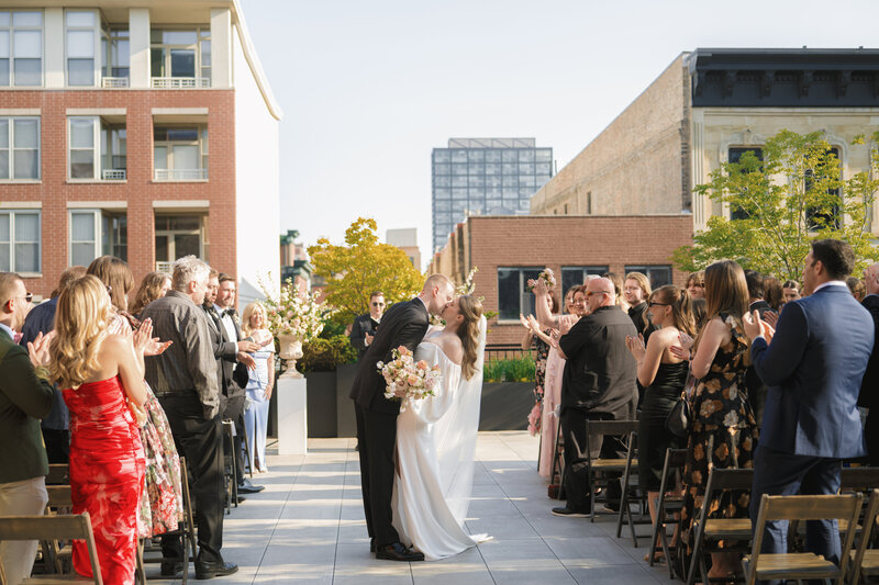 A bride with a long veil and groom in a black suuit smile for the camera near a forest