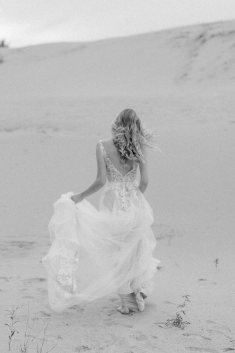 A bride in a lace dress walks on sand dunes in a serene bridal photoshoot.