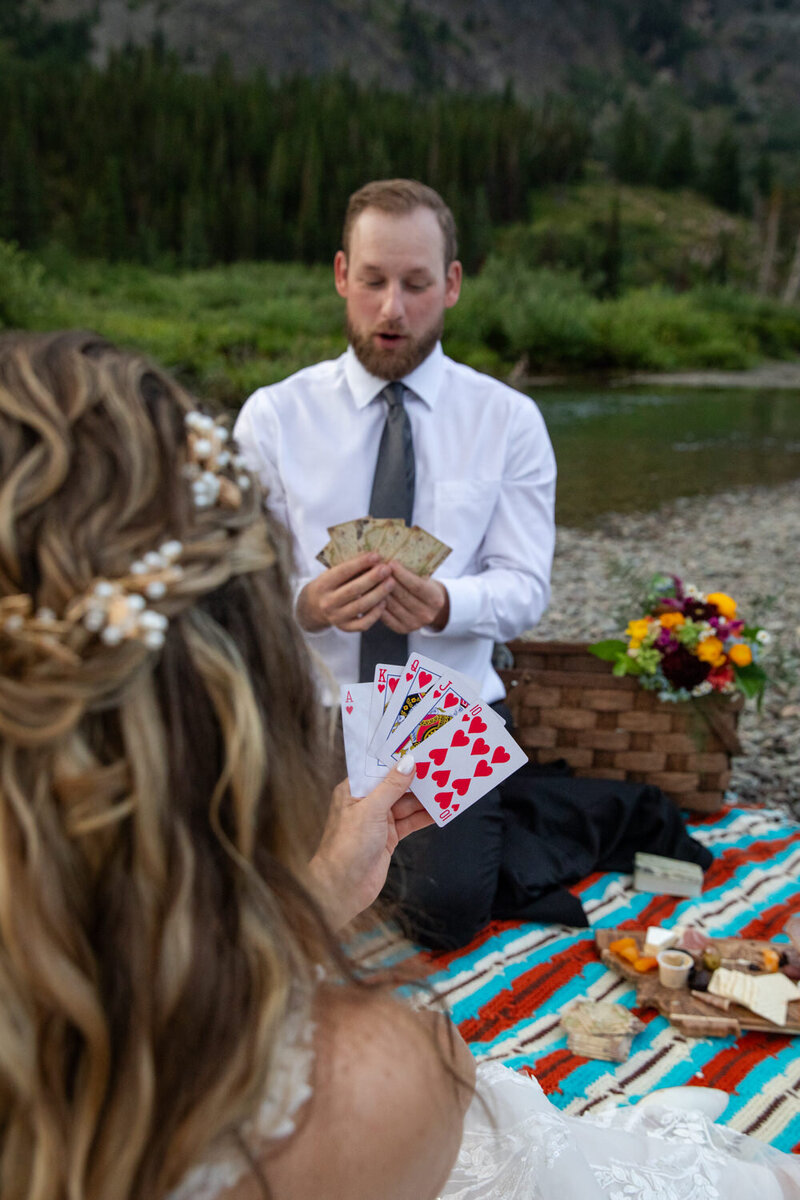 Bride and groom play cards during a riverside picnic, with a colorful blanket, charcuterie, and wildflowers.