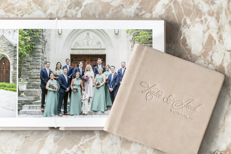 A wedding photo album open to a page showing a wedding party posing in front of a church. Bridesmaids in green dresses, groomsmen in blue suits, and the couple are all smiling. Another album lies closed beside it on a marble surface.