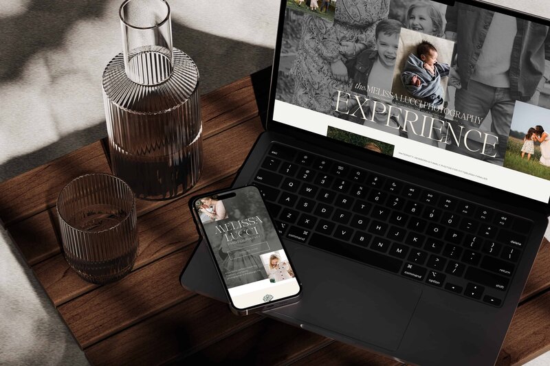 A laptop sits open next to a decanter and glass of water on a dark wood table with an iPhone laying across it showing the beautiful brand and web design for Melissa Lucci Photography crafted by expert Showit Designer and Brand Designer Bayou Brand House.