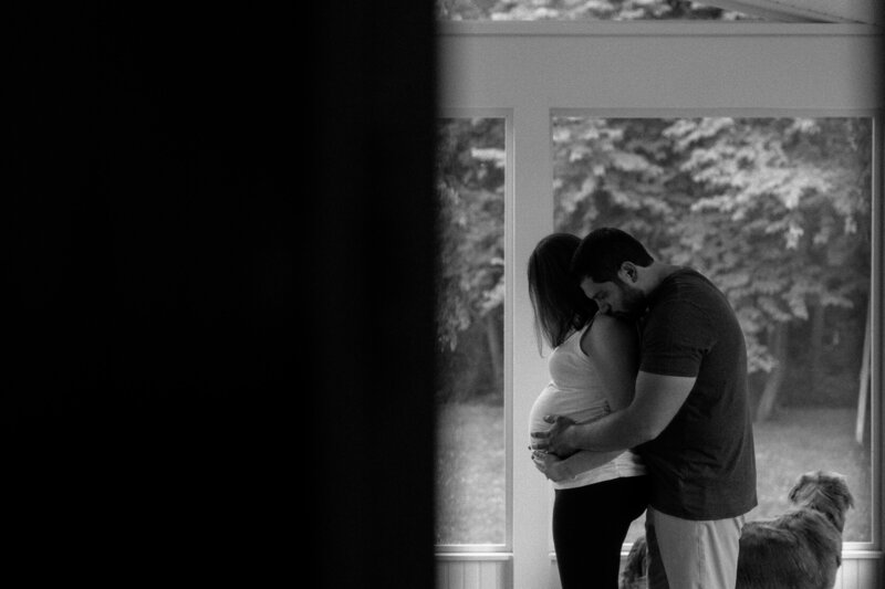 A couple standing in an enclosed porch, with the partner embracing the pregnant woman from behind and a dog looking out the window.