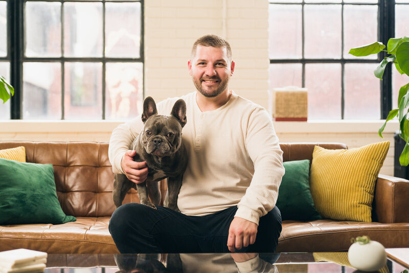Canadian French Bulldog Breeder Peter Kramm is sitting on a couch with his male Blue French Bulldog. Peter is wearing a cream colored ong sleeved shirt he has a short hair cut and a short beared. The blue french bulldog is standing on his right thigh looking at the camera.