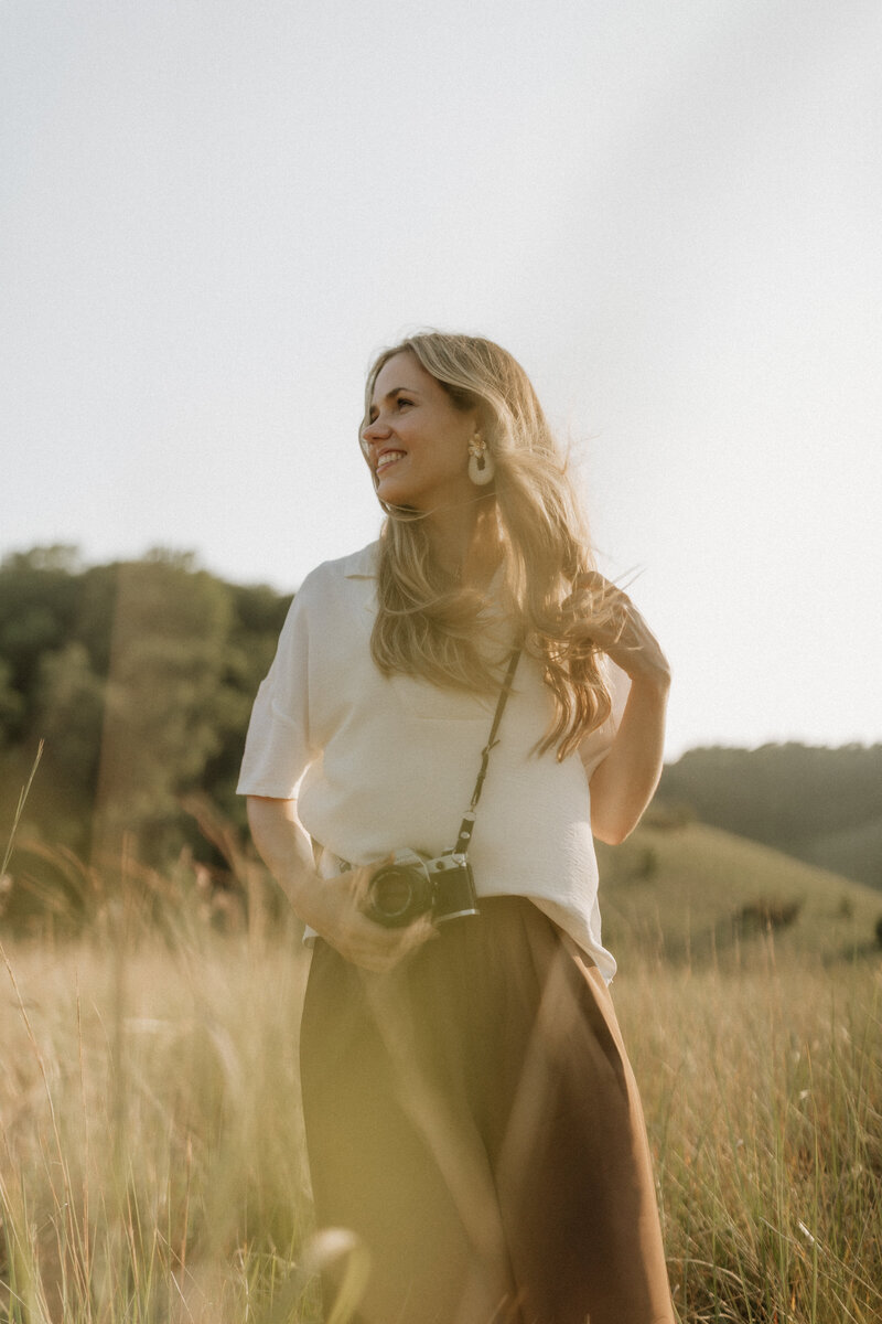 Kaity from Rainstorm Photo & Video standing in a sunlit field with a camera, photographed in warm golden light to capture her creative and joyful spirit as a wedding photographer.