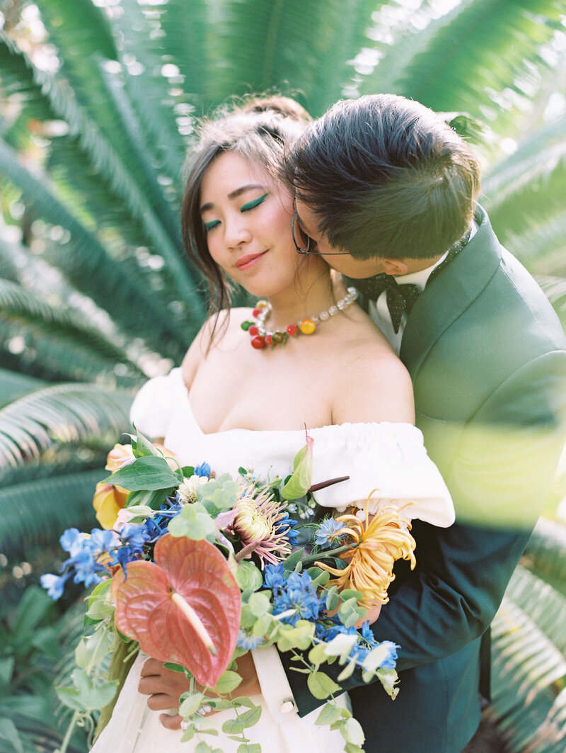 Film photo of a groom kissing his bride on the cheek as she holds her bouquet with exotic tropical flowers in front of tropical plants at Dos Pueblos Orchid Farm in Santa Barbara. 
