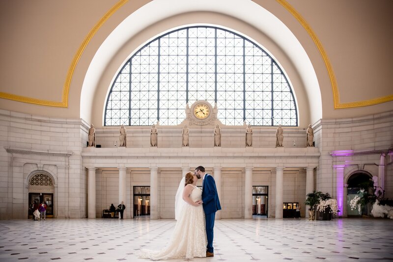 a couple kisses in the middle of a train station on their wedding day. 