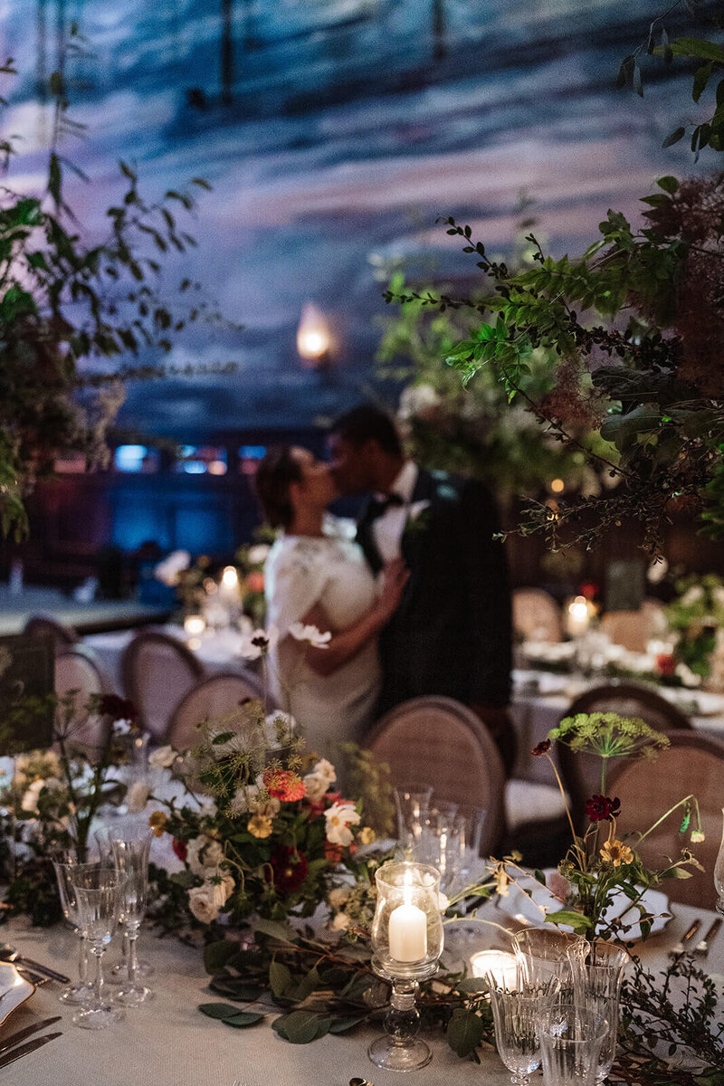 bride and groom stand in the middle of their wedding dinner room and kiss the room is decorated with trees and candles and is in the nomad hotel london