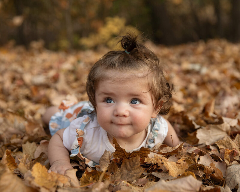 6 month old girl with lots of brown hair and big blue eyes laying in the fall leaves.