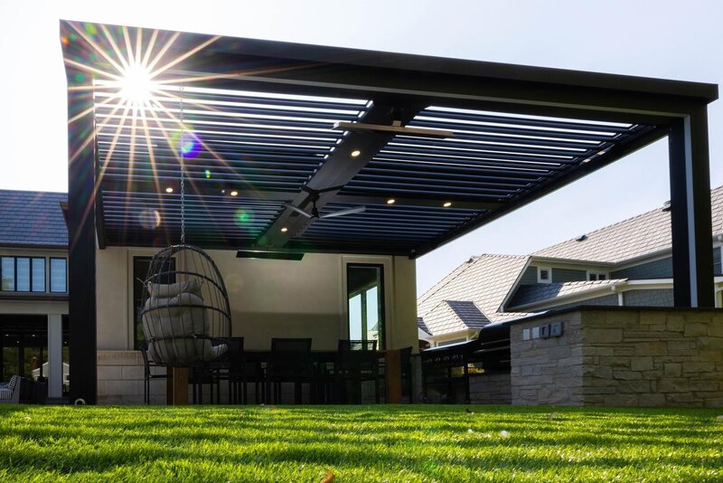 Sun shining through a black StruXure pergola with a hanging chair and dining furniture on a green lawn. 