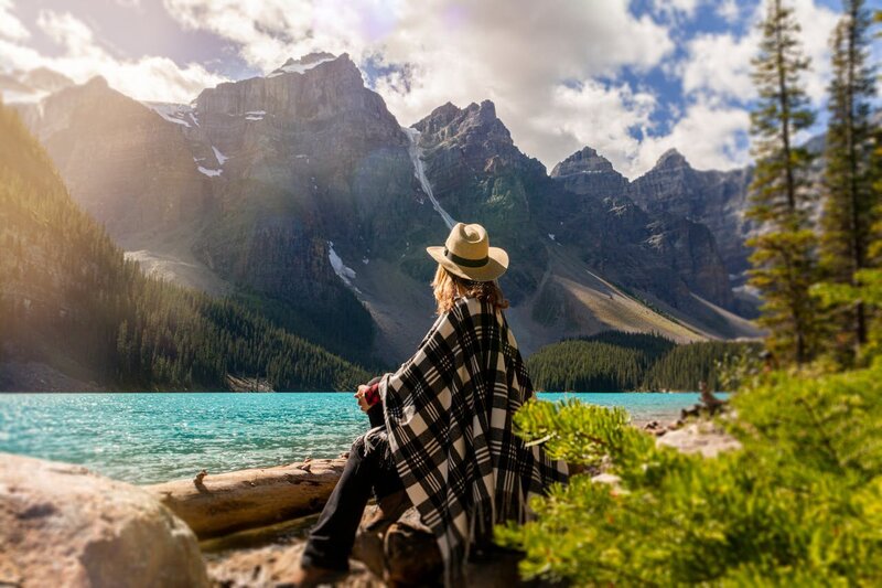 A woman wearing a hat and shall sitting on a rock looking at the mountains and a lake.