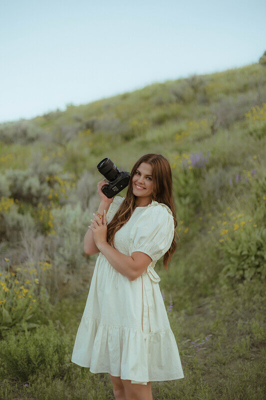 Brand portrait of Brittney Ann Photography. Brittney stands on a hill of sagebrush and greenery, smiling with her camera.