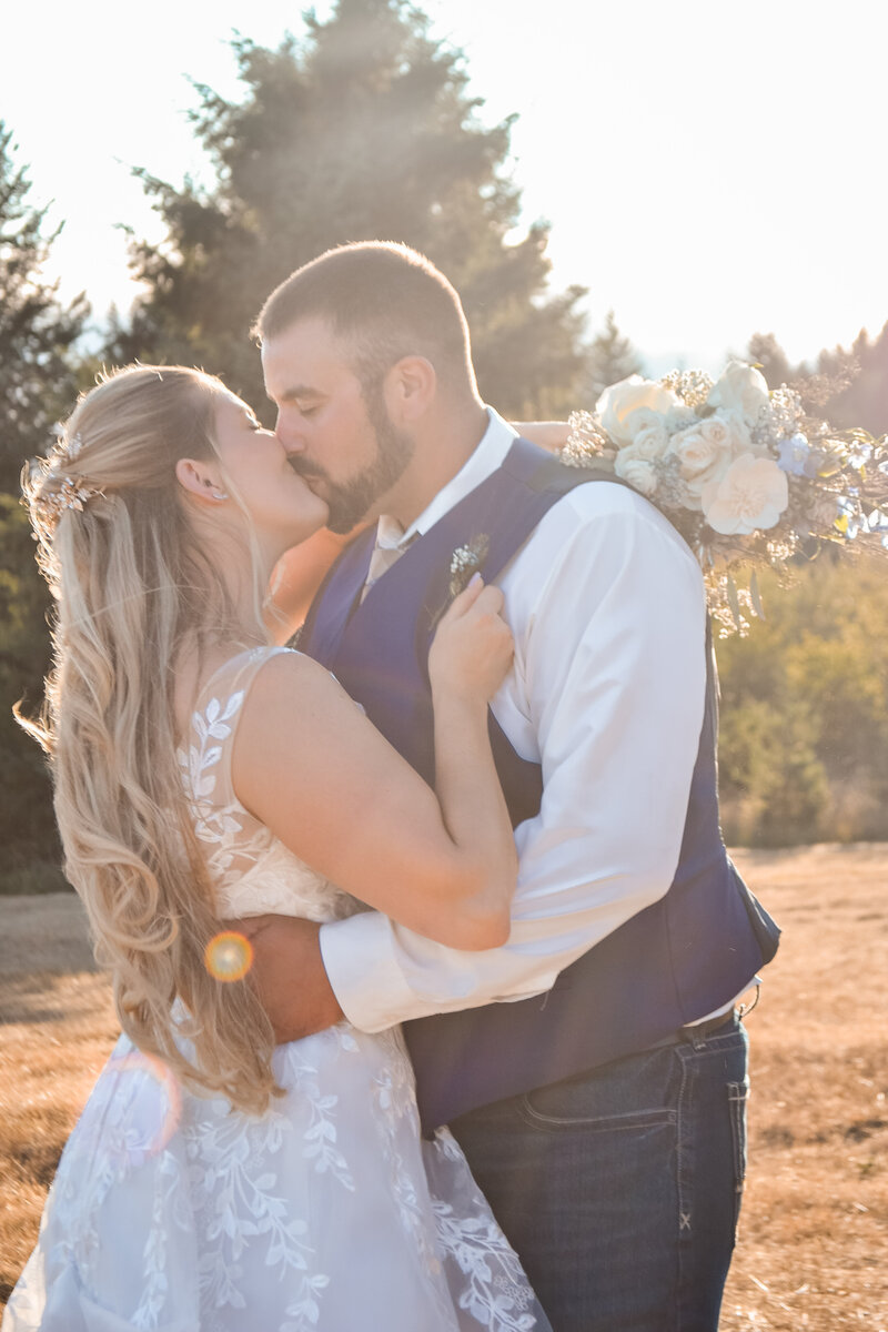 Captivating photo of a bride and groom sharing a kiss at sunset in Salem, Oregon. The bride, holding a beautiful bouquet of white and blue flowers, embraces her groom in this picturesque outdoor wedding moment.