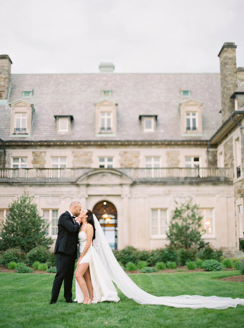 Rhode Island Wedding Photographer | A couple embraces lovingly on a manicured lawn in front of a grand, elegant stone mansion. The bride's long veil flows behind her.