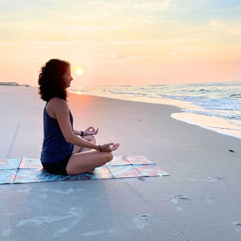Rachel Cline sitting cross-legged in meditation on a beach at sunset