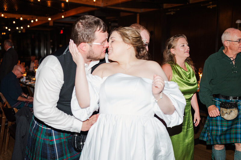 Jewish bride kissing Scottish groom as he holds her from behind on the dance floor in the ballroom at District Winery in DC surrounded by wedding guests