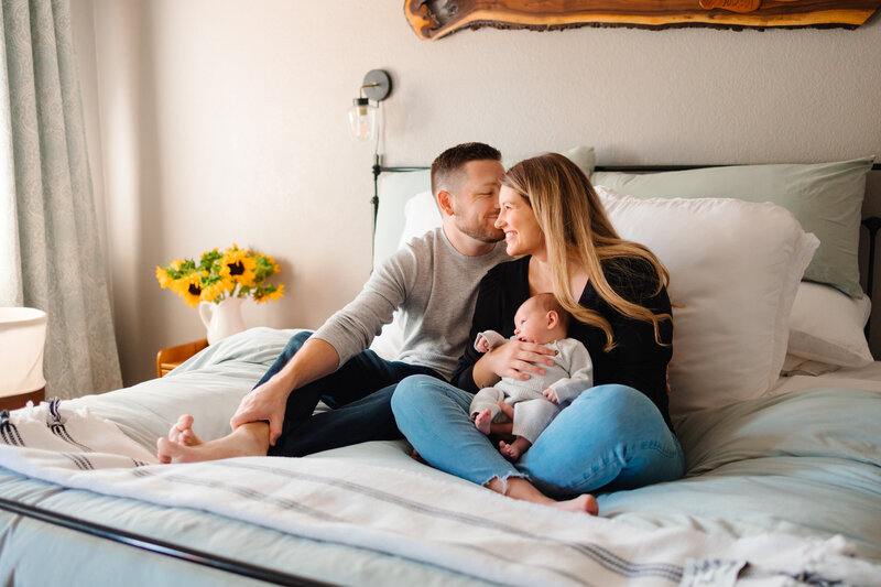 Family photo of two parents holding their newborn baby. The mom is sitting cross legged on their bed and holding their baby while dad sits next to her and kisses her temple. Mom is looking out the window and smiling.