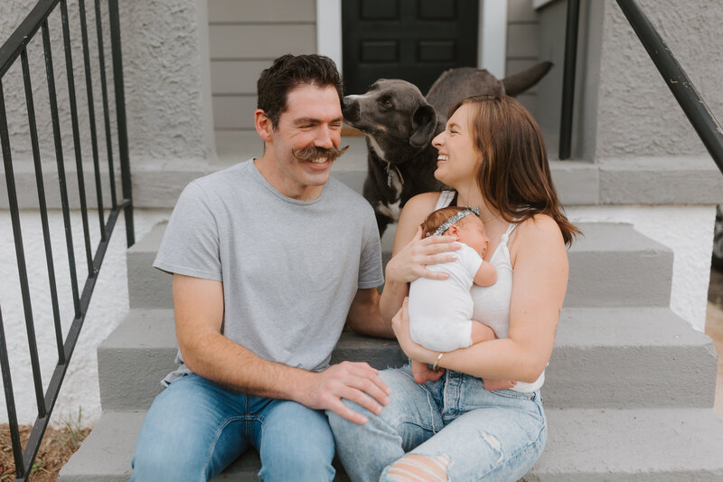 New parents smiling while holding newborn baby with their dog