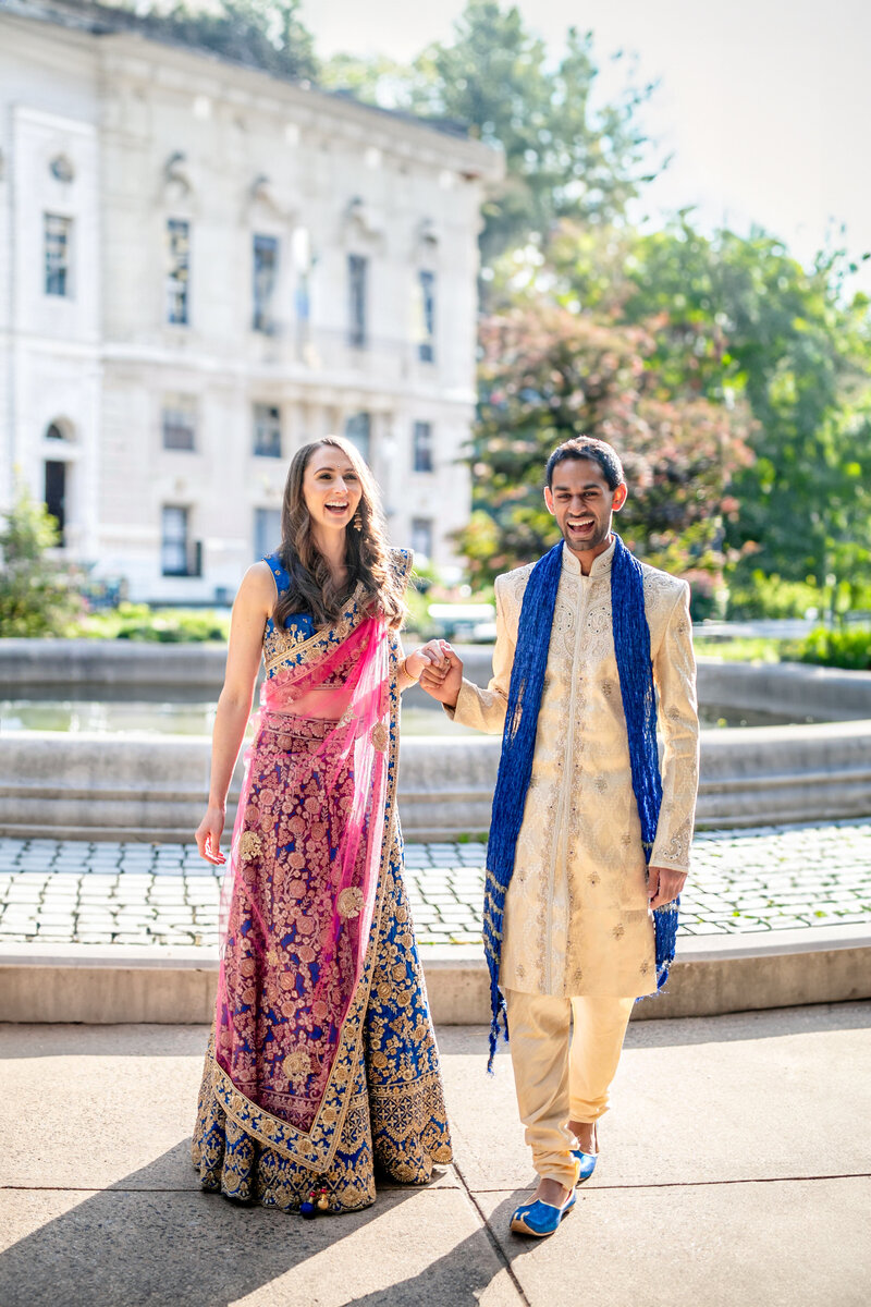 A couple walks hand in hand outdoors, both smiling. The woman wears a vibrant pink and gold traditional Indian outfit, while the man wears an intricate beige outfit with a blue scarf. A large building and greenery are in the background.