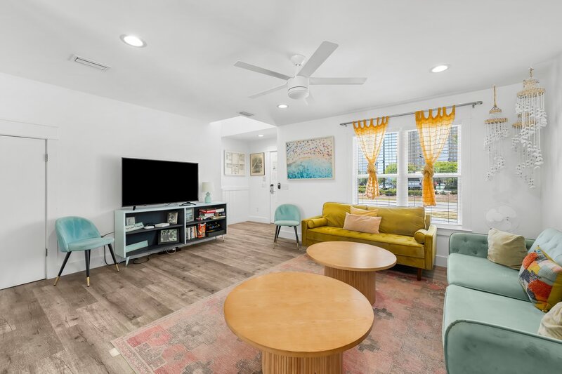 Bright and inviting living room with mid-century modern furniture, large windows with gold sheer curtains, and beach-inspired art.