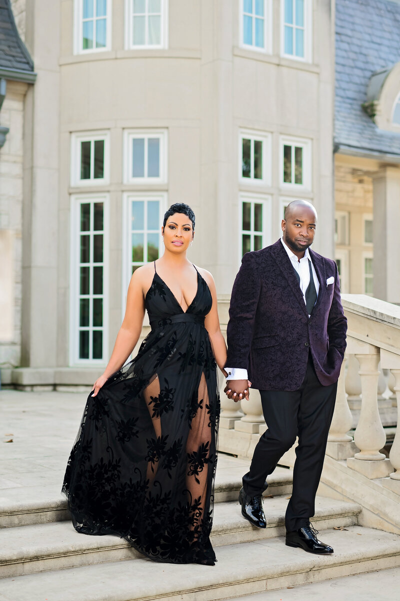 A couple walks down a grand outdoor staircase in front of a manor. The woman wears a flowing black lace down, and the man is in a dark velvet suit.