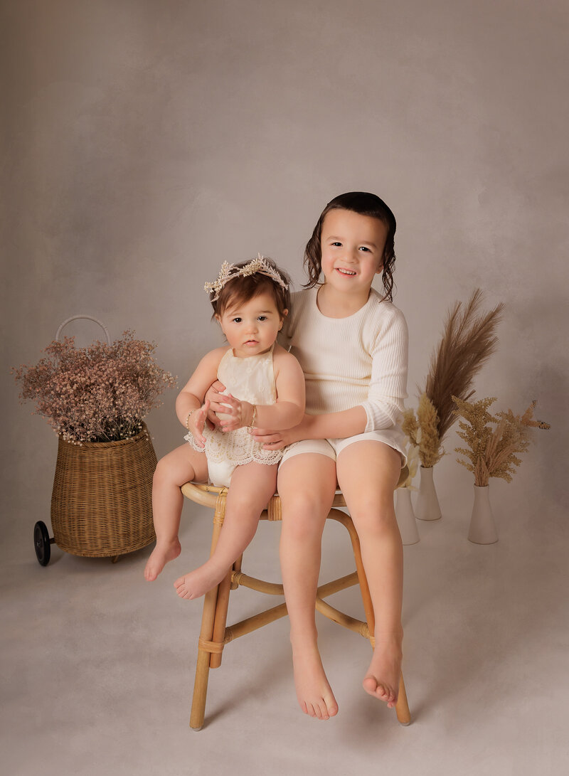Big brother sits with his little sister on a chair for her first birthday photoshoot in brooklyn, ny. Both are smiling at the camera. Captured by premier Brooklyn NY family photographer Chaya Bornstein Photography.