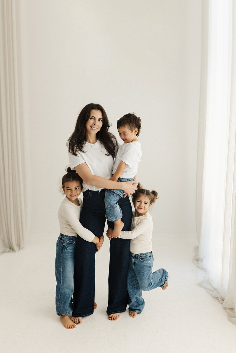 A mother and her three child smiling at the camera wearing jeans and white tops in a 90s inspired family photoshoot in the christchurch family photography studio by lucy jane photography