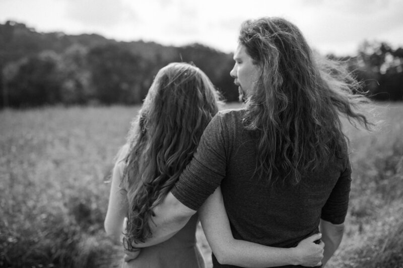 A couple stands together, close in a field, embracing with long, flowing hair gently blowing in the breeze under a cloudy sky.
