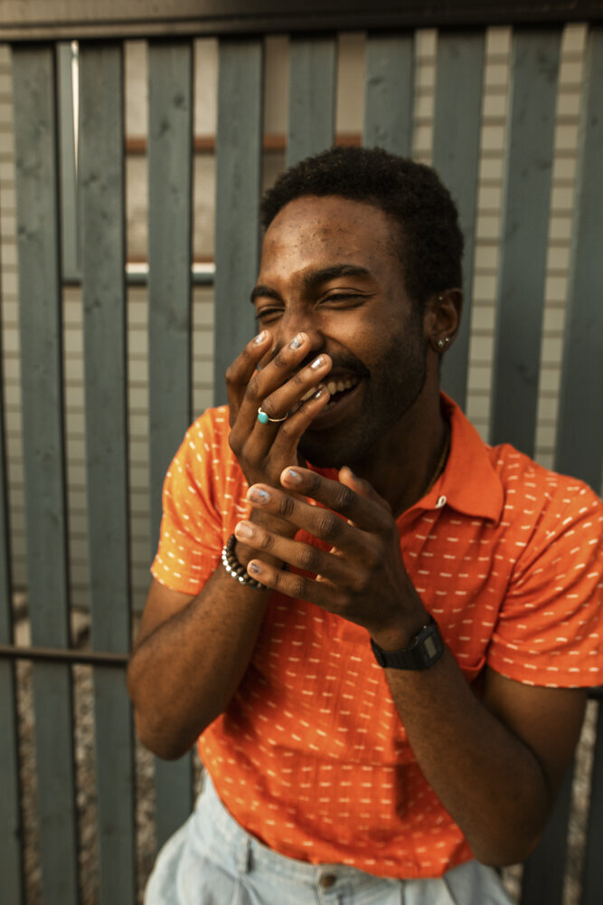 A masculine-presenting person of color stands outside against a fence, laughing while bringing their hands up to their face. They are wearing silver nail polish, a ring, and multiple bracelets, as well as a bright orange polo.