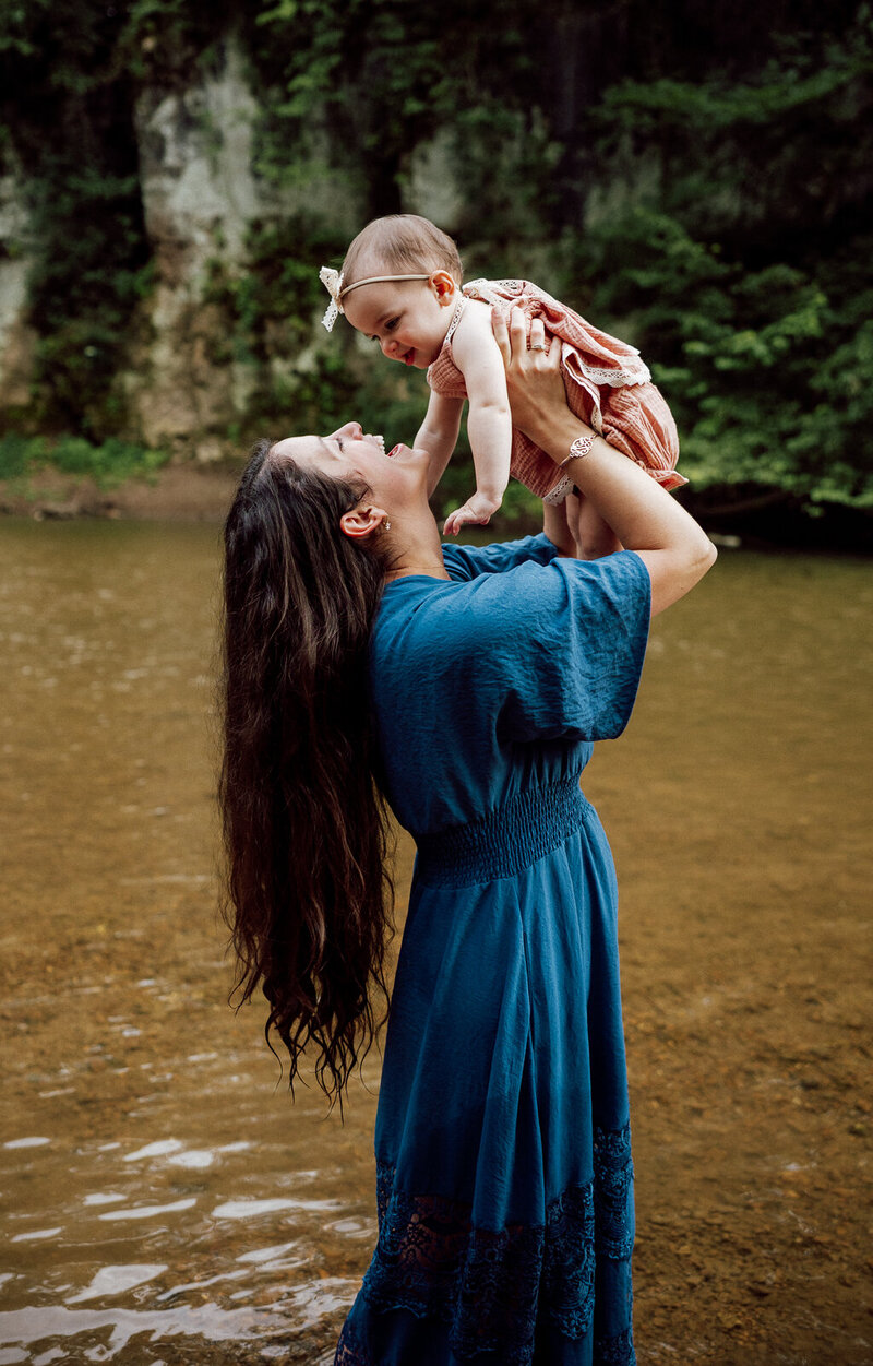 Mother standing at the creek holding one year old daughter in the air smiling at one another. 