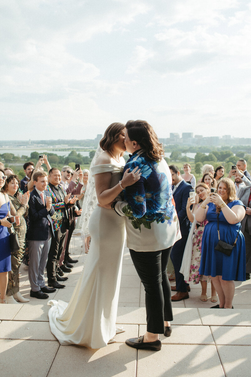 lesbian brides kissing on the terrace overlooking DC surrounded by wedding guests