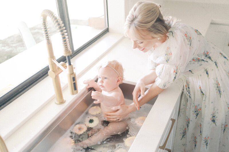 mom in white dress giving her baby girl a bath in the kitchen sink.