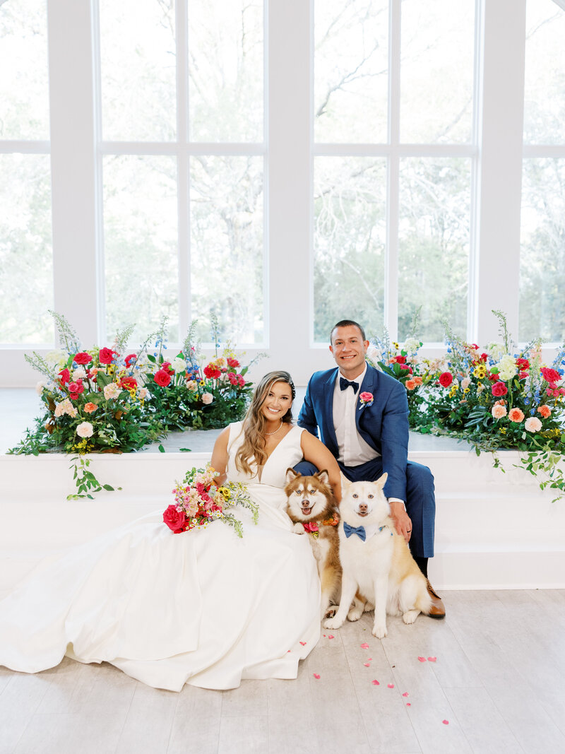 bride and groom sitting together with their two dogs surrounded by pink, yellow, and blue florals