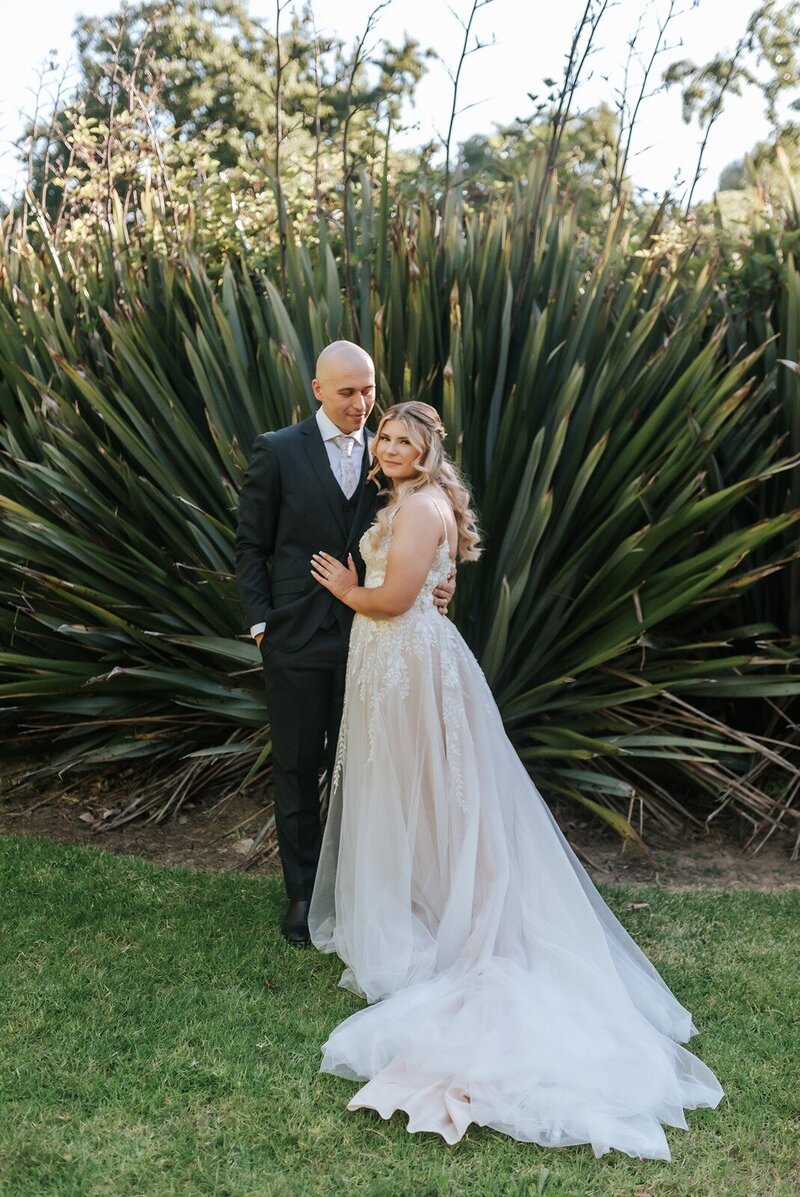 A bride and groom standing in front of a large bush for wedding photos