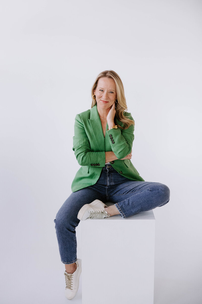 Heather Frick executive coach in a green blazer and jeans sitting on a white square stool