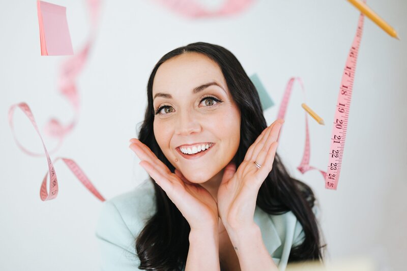 Woman in pastel blue blazer frames her face and smiles for the camera as pencils, post its and measuring tapes are suspended above her.