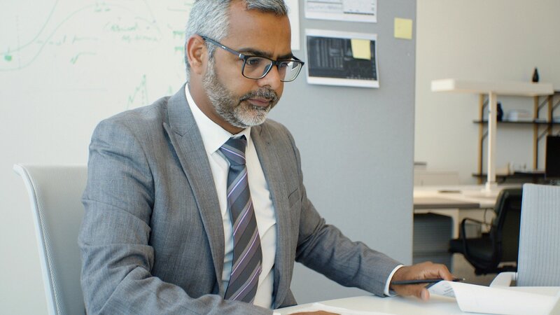 A middle-aged man with medium skin tone, in a suit with a gray jacket and purple and gray striped tie, at a desk