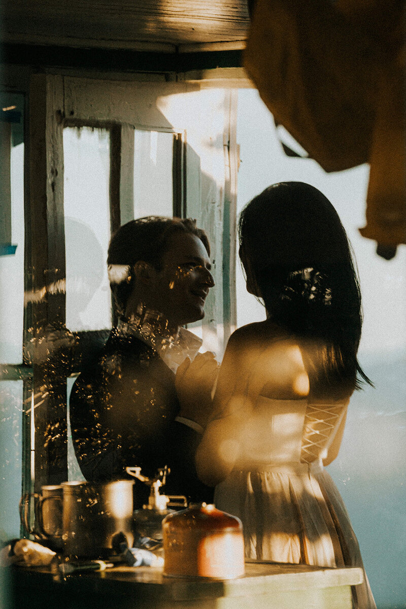 their adventure elopement photographers shoot through the glass of a fire lookout to capture this couple embrace in their wedding attire. 