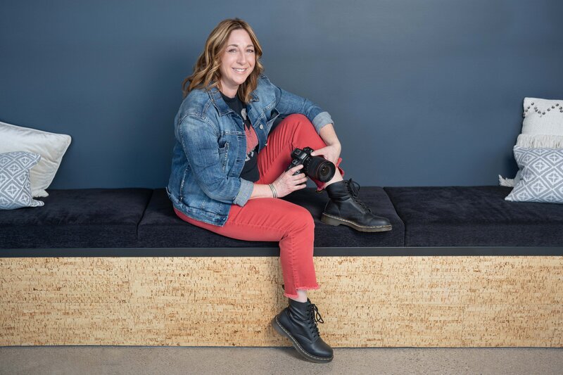 A female photograper is seated on a bench in a casual pose. She is holding a Canon camera and wearing red pants, Doc Marten boots, and a denim jacket over a Red Hot Chili Peppers tshirt