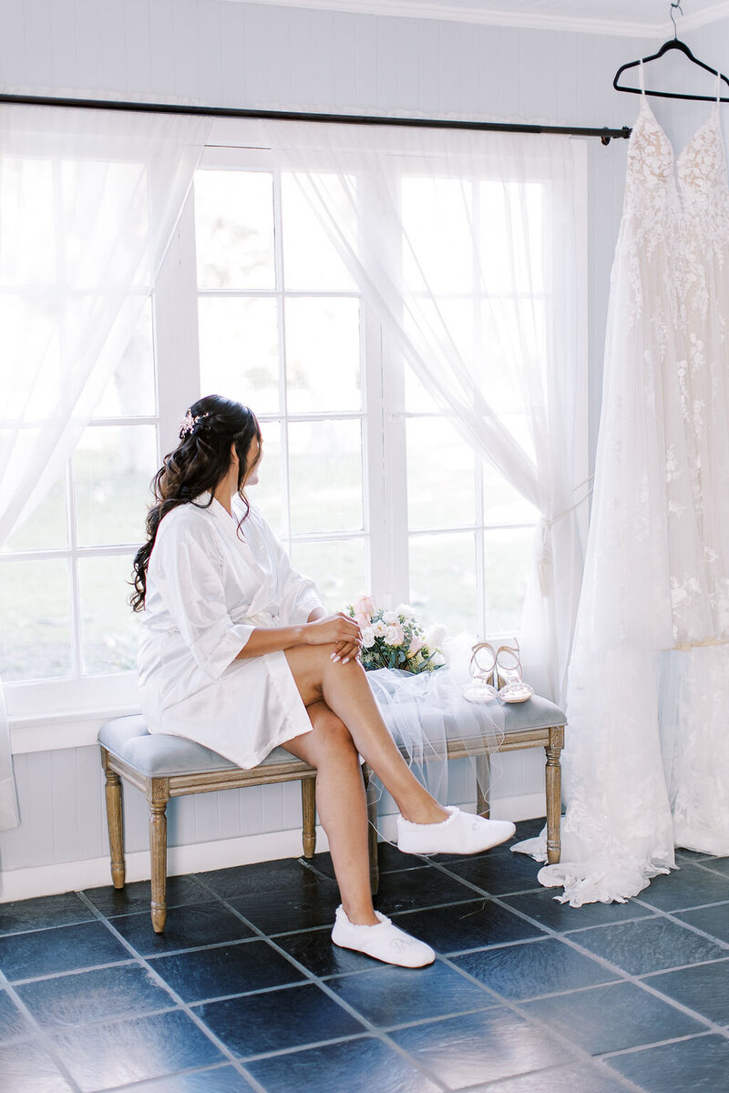1-radiant-love-events-bride-with-robe-on-sitting-by-window-looking-outside-wedding-dress-hanging-romantic-elegant-timeless