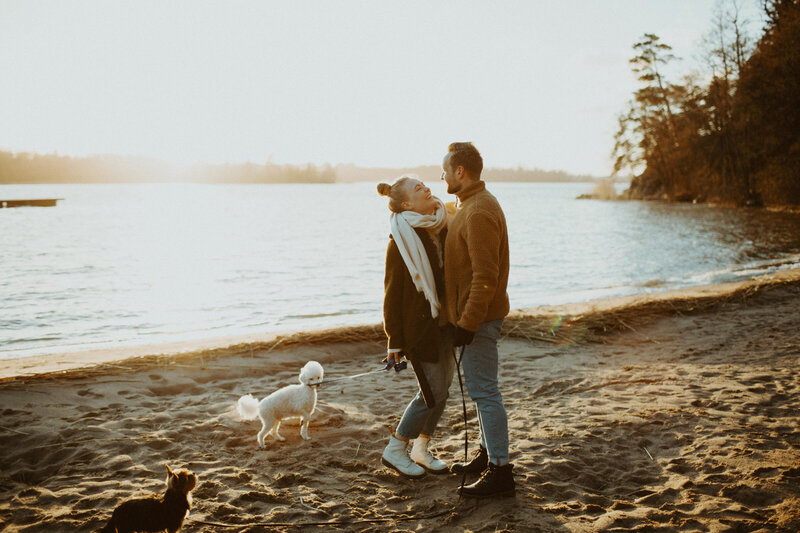 Kihlapari seisoo rannalla lähekkäin ja hymyilee toisilleen rannalla katsoen toisiinsa Kivenlahdessa Espoossa
