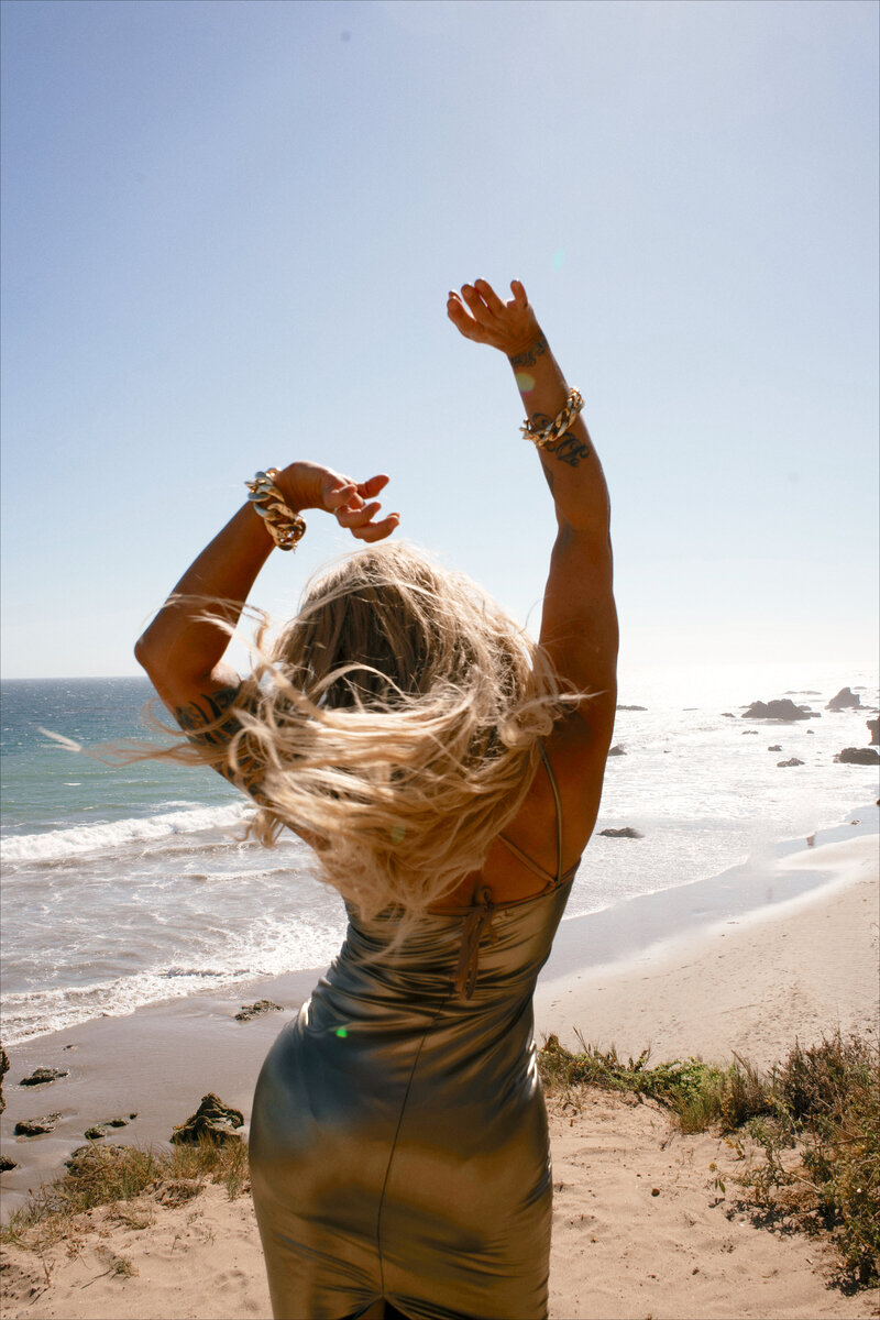 Back view of Nicky Lynn with arms raised on a rocky cliff, symbolizing letting go and self-liberation