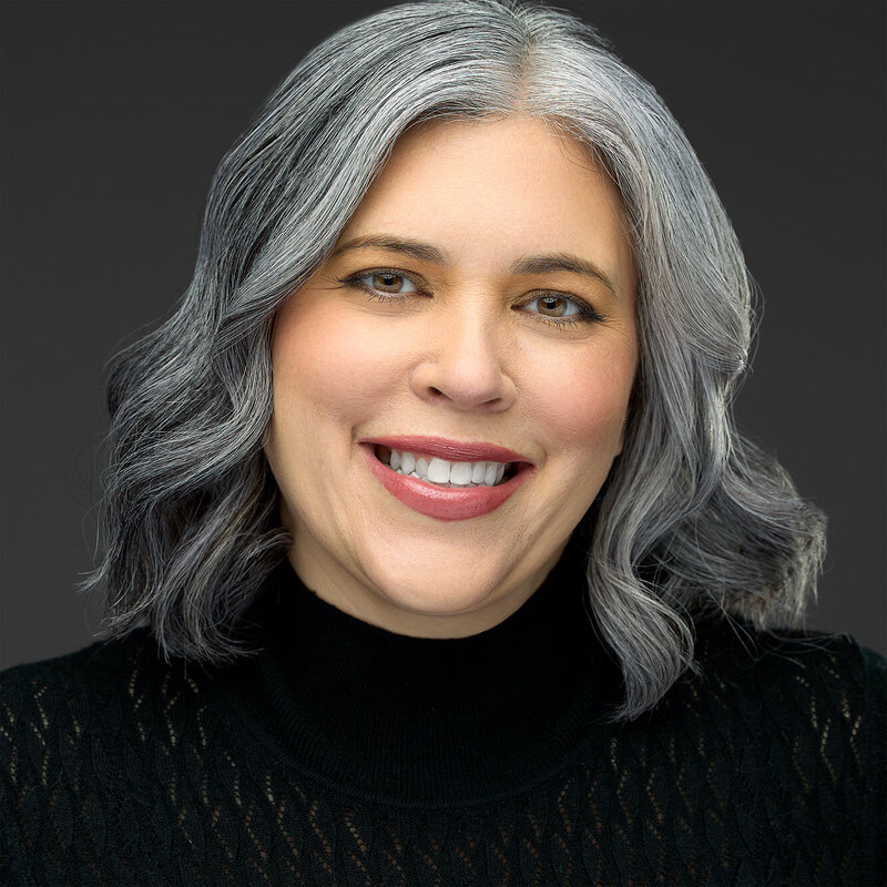 A close-up portrait of a woman with shoulder-length gray hair, smiling at the camera. She is wearing a black sweater, and the background is a solid dark color, creating a sharp contrast with her hair and attire.