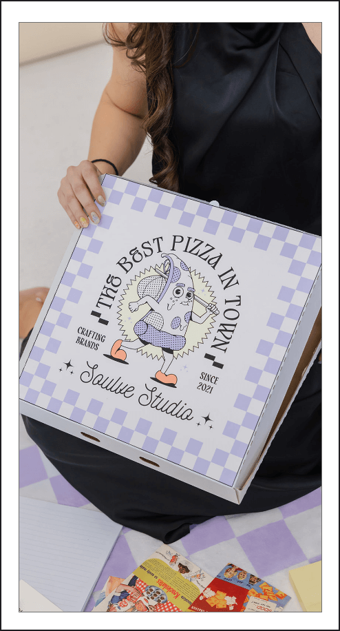 A woman holds a custom pizza box designed by Soulve Studio, featuring a playful illustration of a pizza slice character and the slogan "The Best Pizza in Town." The box has a checkered purple and white pattern with the Soulve Studio logo and a vintage-inspired aesthetic. The design reflects the studio's creative approach to branding and packaging, blending fun and nostalgia with a modern twist.